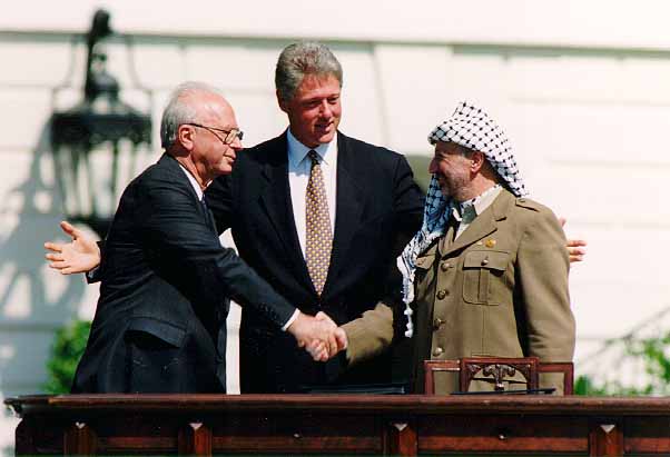 Three men stand outside shaking hands, with a third smiling and extending his arms, symbolizing understanding amid the Israeli-Palestinian Conflict.