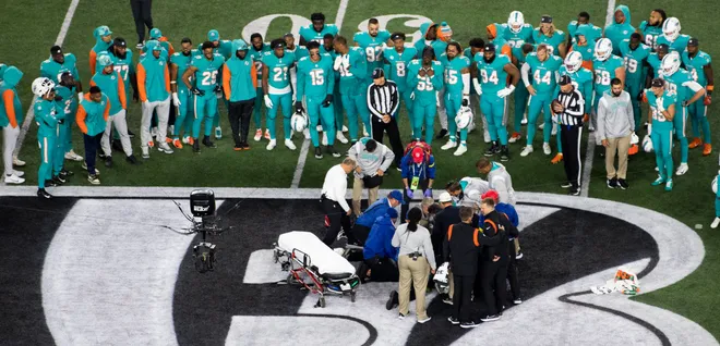 Medical staff assist an injured NFL player on the field as teammates and officials stand in a circle around them near the midfield logo.