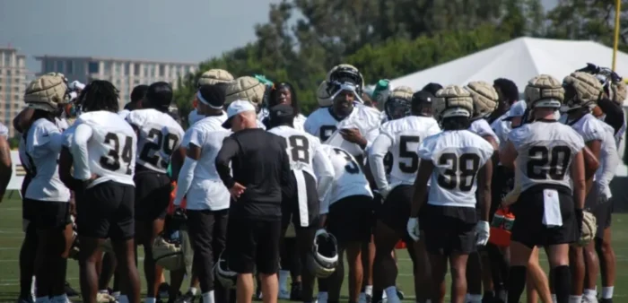 A football team in practice jerseys and helmets gathers in a huddle on a grassy field, with a coach addressing them.