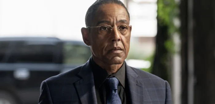 A man in a dark plaid suit, glasses, and tie stands outside near a blurred vehicle, looking ahead with a serious expression.