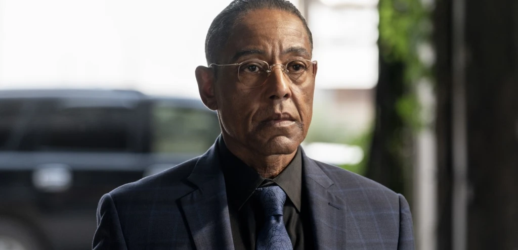 A man in a dark plaid suit, glasses, and tie stands outside near a blurred vehicle, looking ahead with a serious expression.