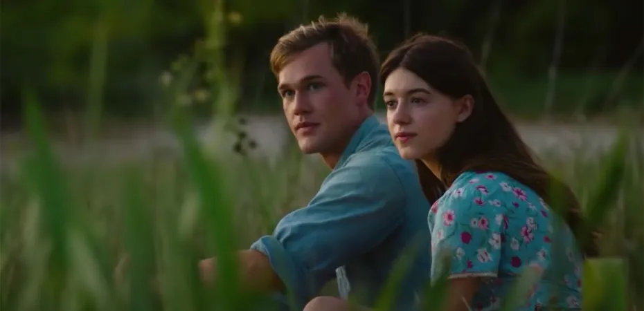 A young man and woman sit together in tall grass, looking into the distance, with greenery in the background.