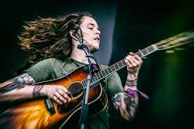 Person with long hair playing an acoustic guitar on stage, with tattoos on their arms and wearing a green shirt.
