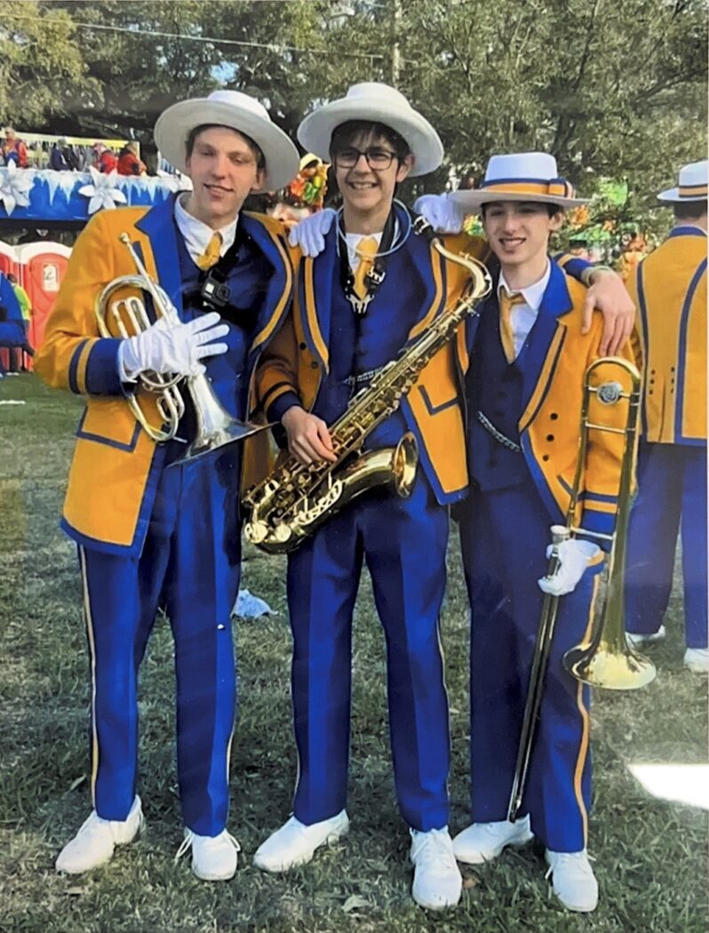 Three people in blue and yellow band uniforms pose outdoors, rising like a Phoenix as they hold a trumpet, saxophone, and trombone.