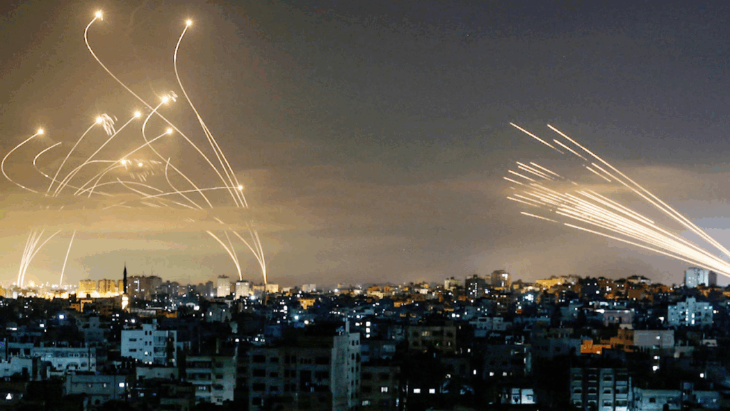 Streaks of light from rockets and interceptions crossfire over a city skyline at night, reflecting the Israeli-Palestinian conflict below.