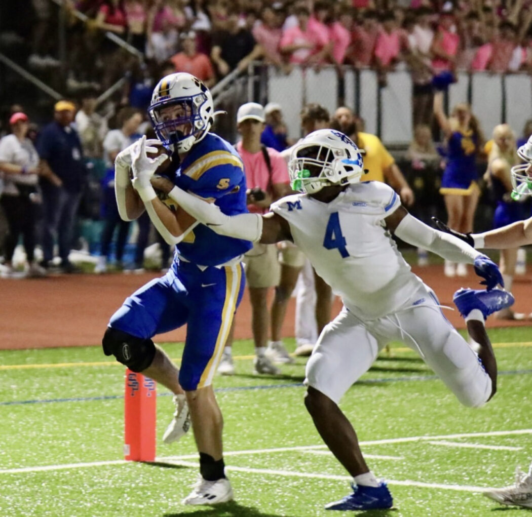 A football player in blue runs with the ball as a Saint Paul's Football Wolves defender in white tries to tackle him near the end zone at night.
