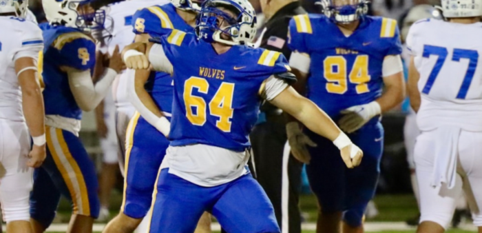 A football player in a blue Mandeville High School Wolves uniform, number 64, stands with arms outstretched on the field among other players.