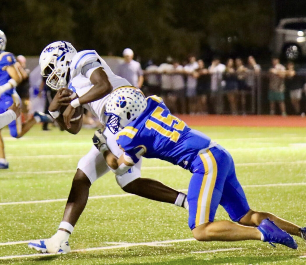 A Saint Paul's Football player in white is tackled by a Mandeville High School player in blue and yellow on a lit field with spectators watching.