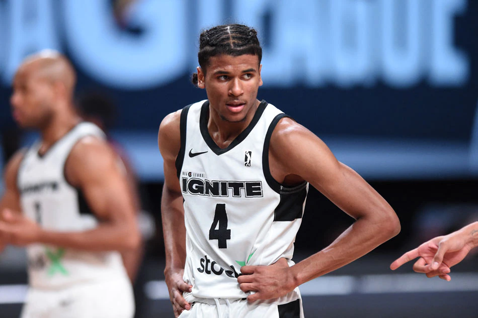 A young basketball player in a white and black G League Ignite jersey stands on the court, with other basketball players in the background.