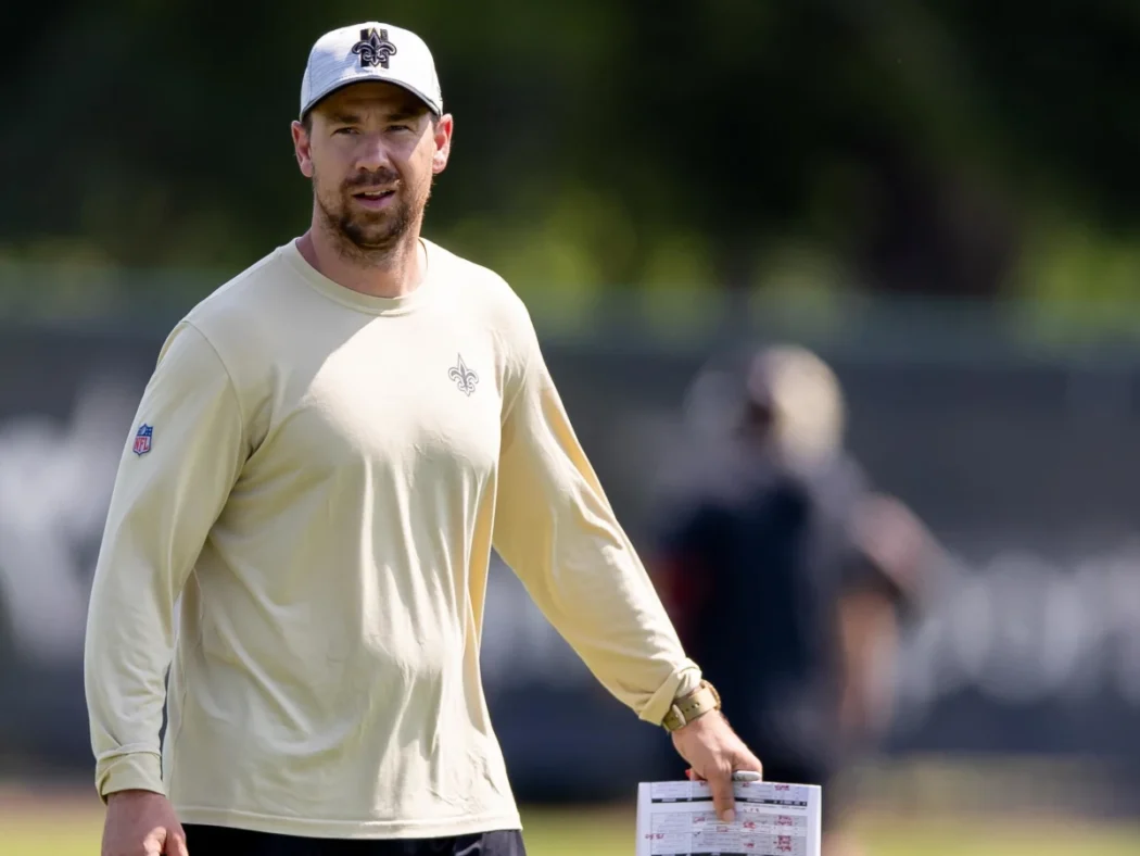 A man in a light-colored New Orleans Saints shirt previews the 2024 season outdoors, holding a sheet of paper with trees in the background.