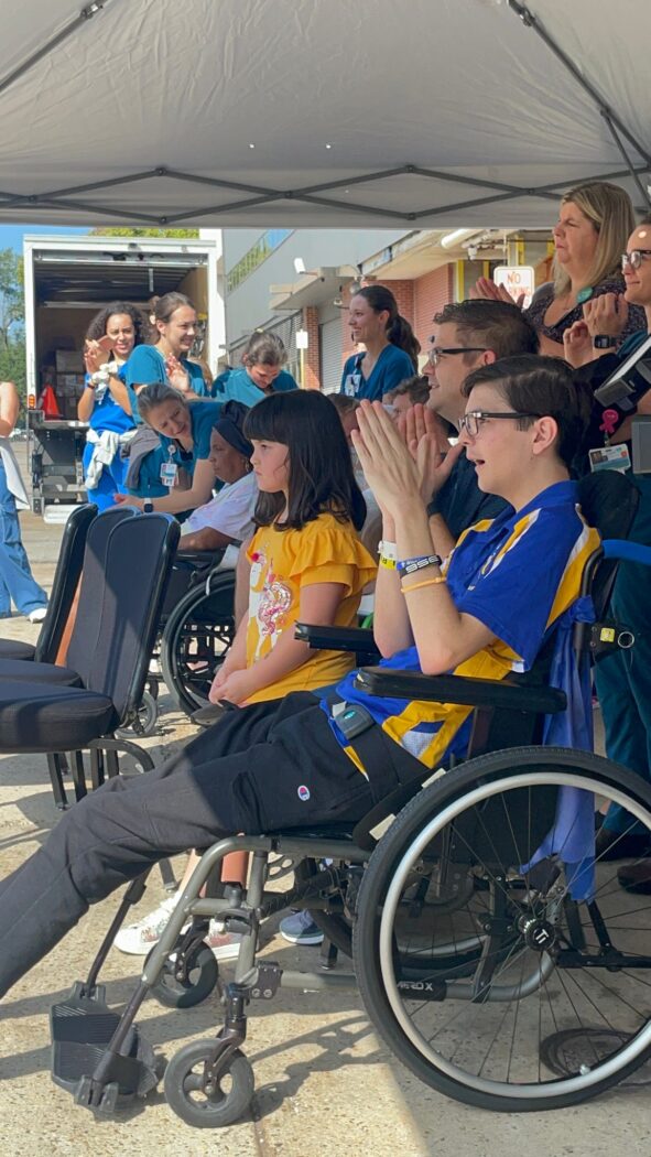 A group of children and adults, including some in wheelchairs, sit under a tent outdoors in Phoenix, watching something unfold.