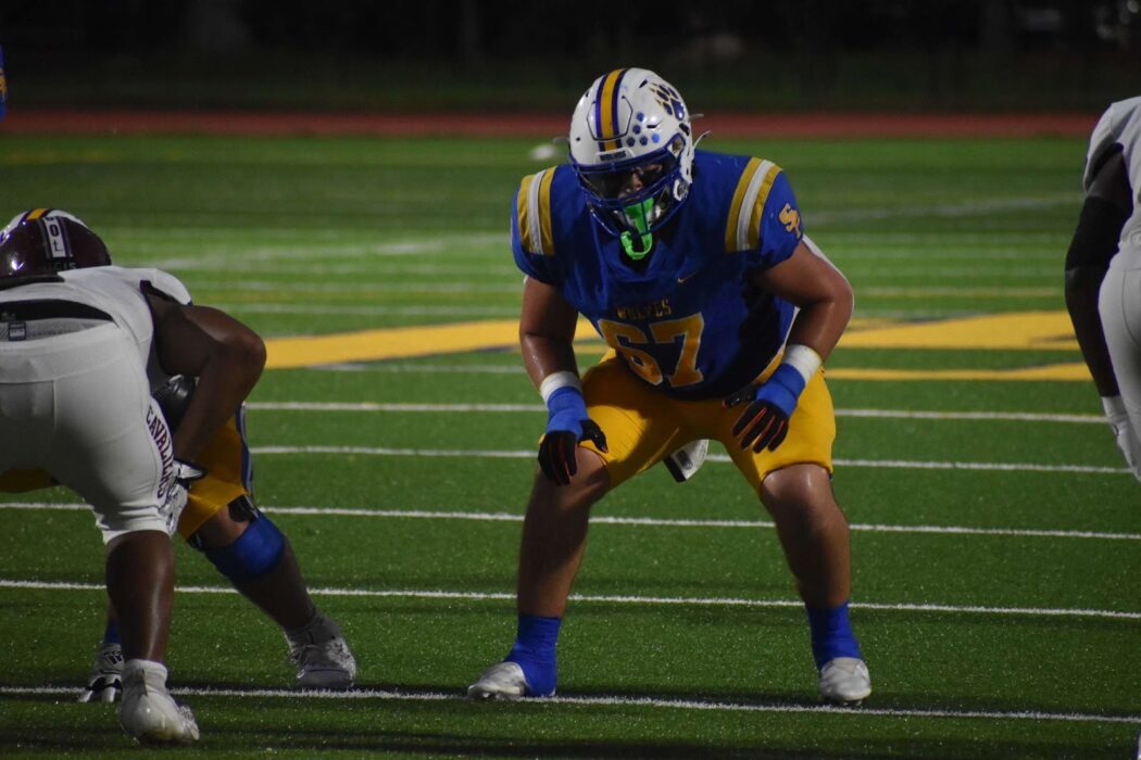 A John F. Kennedy Cougars football player in blue and yellow crouches on the field, facing two Wolves in white uniforms.