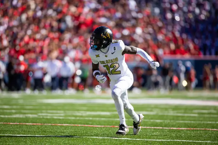 Travis Hunter, in a white Colorado uniform, runs on the field during a game, making waves in the Heisman race as the crowd blurs behind him.