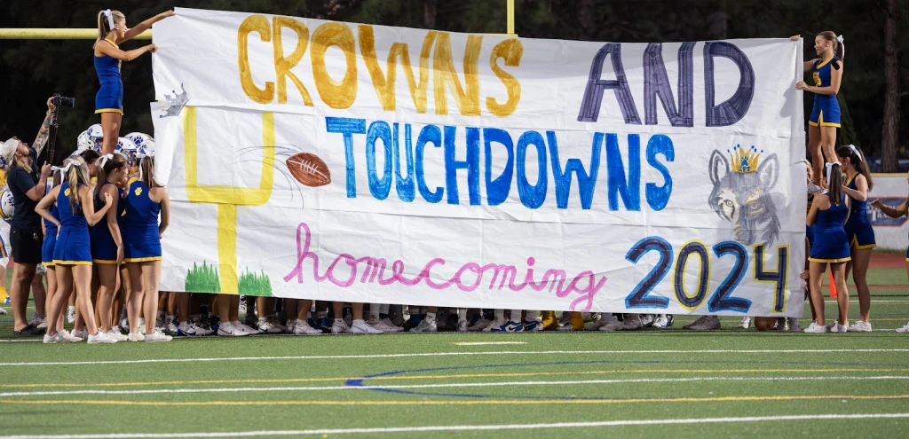 High school cheerleaders hold a "Crowns and Touchdowns Homecoming 2024" banner for the John F. Kennedy Cougars on the football field.