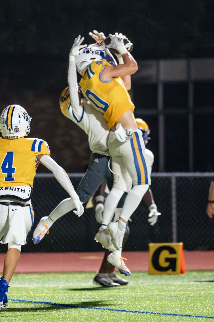 Two John F. Kennedy Cougars in yellow leap to intercept a pass during the Homecoming night game, as a Wolves player looks on.