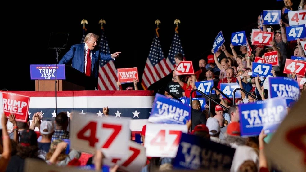 A man speaks at a rally podium with "TRUMP VANCE" signs amid 2024 presidential election buzz; supporters wave "47" signs and flags.