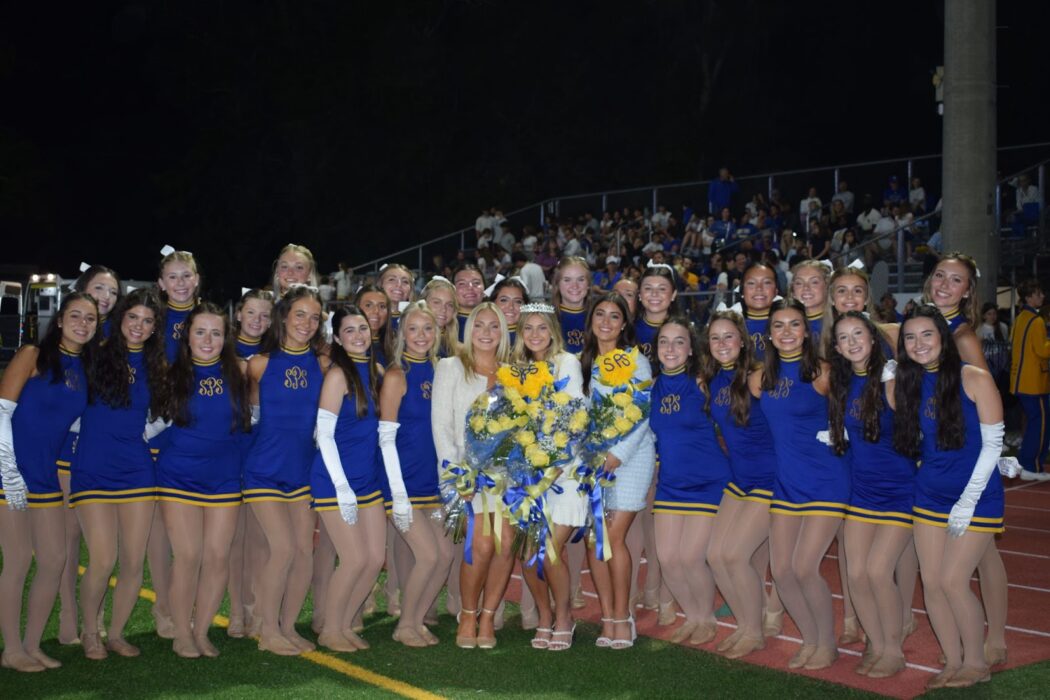 A group of John F. Kennedy Cougars cheerleaders in blue uniforms poses with three holding bouquets on the football field at Homecoming.