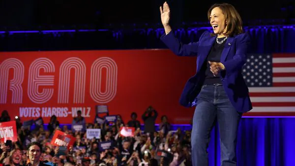 A woman in a blue blazer waves on stage before a crowd, with a U.S. flag, "Reno" sign, and 2024 presidential election energy in the air.
