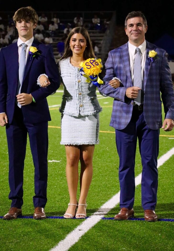 Three people stand arm in arm on a football field; the woman in the center holds a Homecoming bouquet with blue letters.