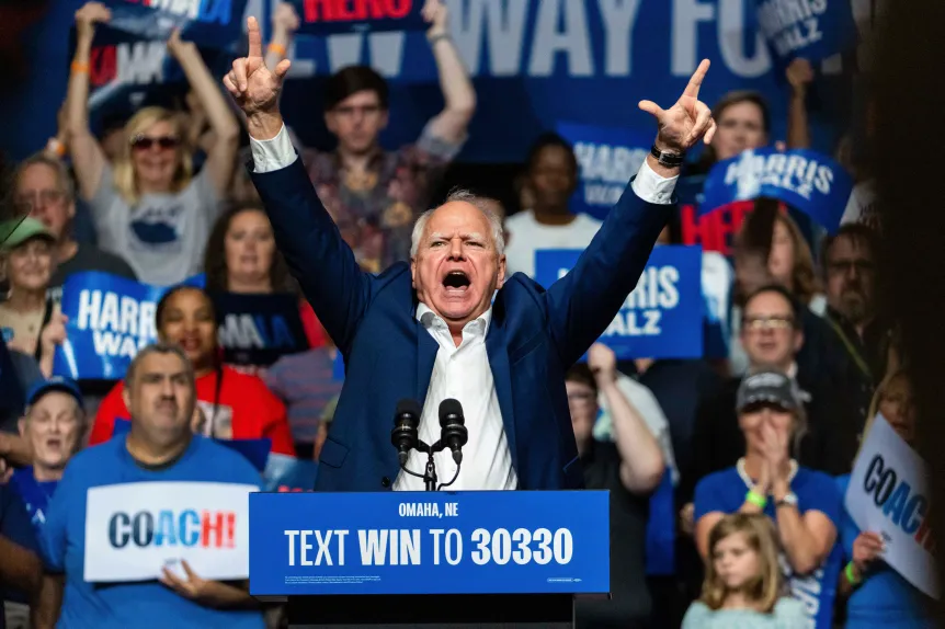 A man stands at a podium with raised arms, rallying a crowd as banners highlight battleground state polling for the 2024 presidential election.