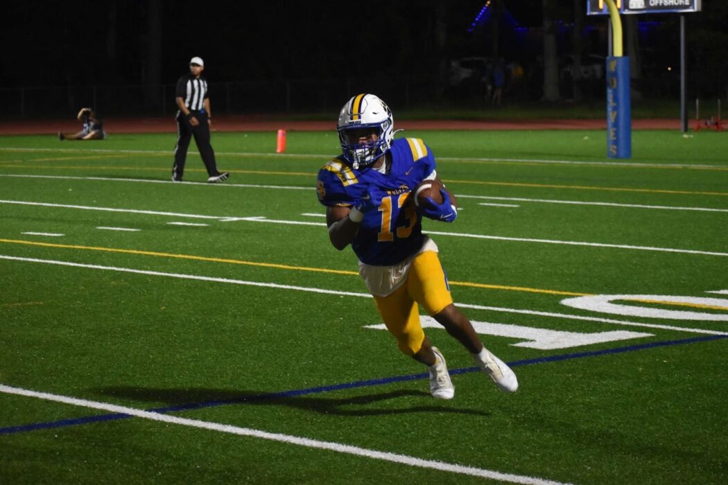 A John F. Kennedy Cougars football player in blue and yellow runs with the ball during a nighttime Homecoming game, referee in the background.