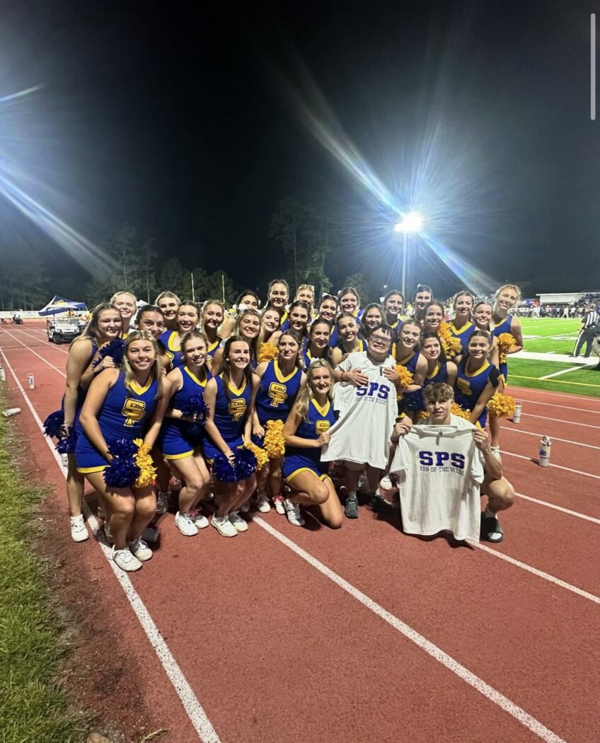 A group of cheerleaders in blue and yellow uniforms pose on a track at night for Homecoming, with two holding SPS shirts in front.