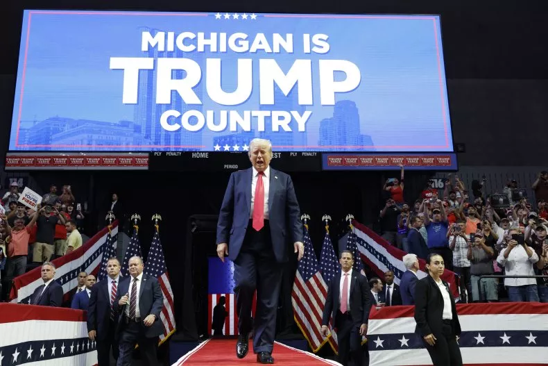 A man in a suit walks on stage at a rally with a "Michigan is Trump Country" sign, reflecting battleground state polling momentum.