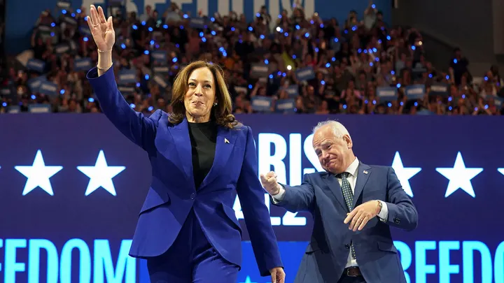 A woman in a blue suit waves on stage with a man, as a crowd holding signs cheers during a 2024 presidential election event.