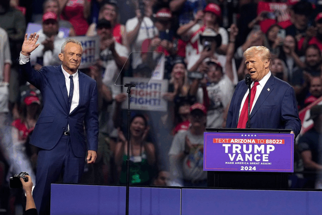 A man in a blue suit waves while another speaks at a "Trump Vance 2024" podium, energizing the crowd ahead of the 2024 presidential election.