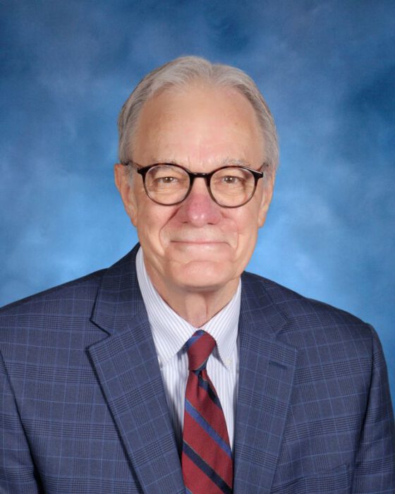 An older man with gray hair and glasses in a blue plaid suit poses, reflecting his dedication to alumni relations, against a blue studio background.