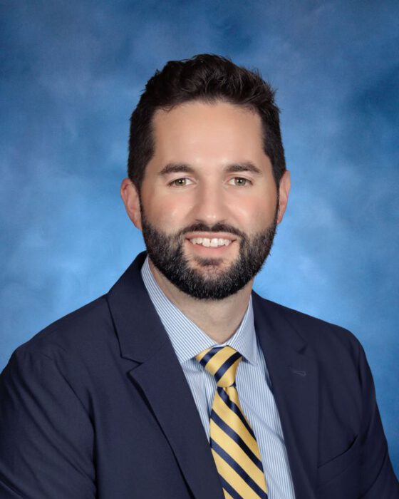 A man with short dark hair and a beard in a navy suit, representing Institutional Advancement, poses before a blue background.